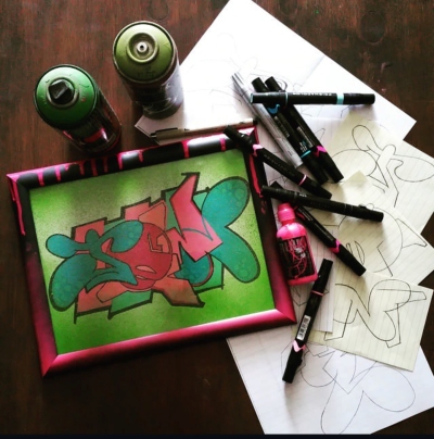 Markers, spray paint, sketches on lined paper, and a green and pink graffiti piece in a black and dripping pink paint frame