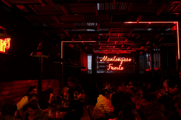 Inside Bar Callejón. A crowd of people gathers, seated at tables under the soft red-orange glow of a neon sign.