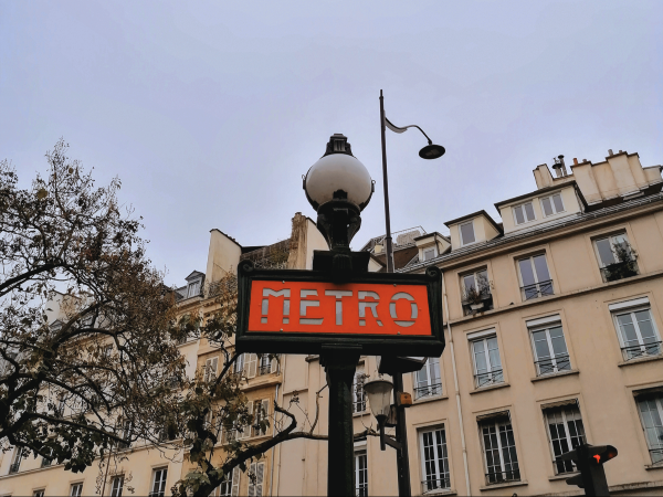 Black lamppost with orange "METRO" sign in front of a tan building in Paris.