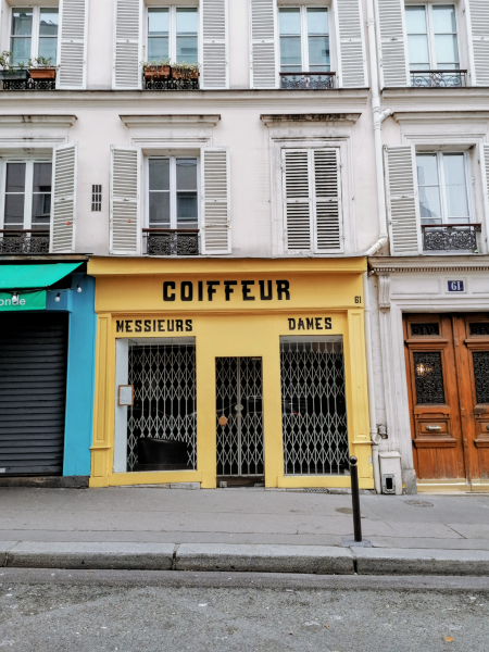 Brightly painted businesses on a Paris street. Centered: "Coiffeur" in black all-caps letters on a yellow background.