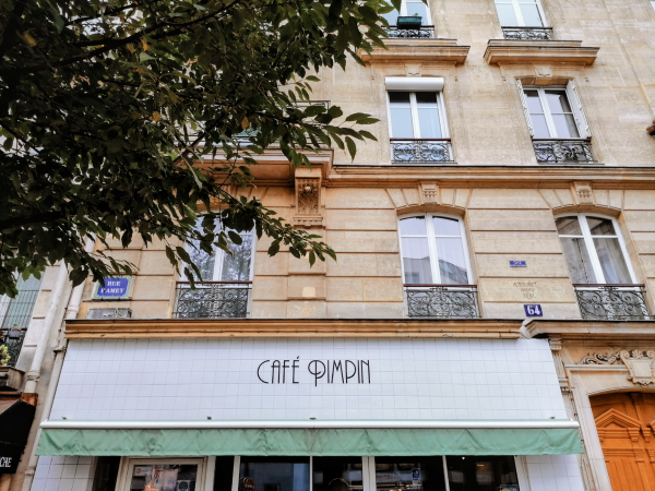 Framed by a leafy tree, "CAFÉ PIMPIN" store sign in black on white tiles, with a thin lime green curtain below.