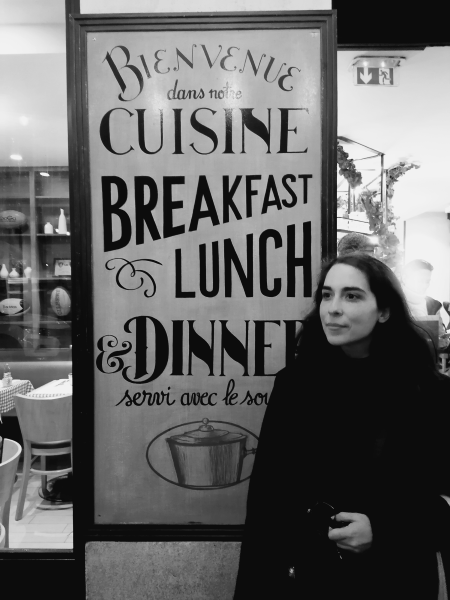 Black and white portrait of Ana Parracho in front of a street advertisement for a restaurant advertising "BREAKFAST, LUNCH, & DINNER."