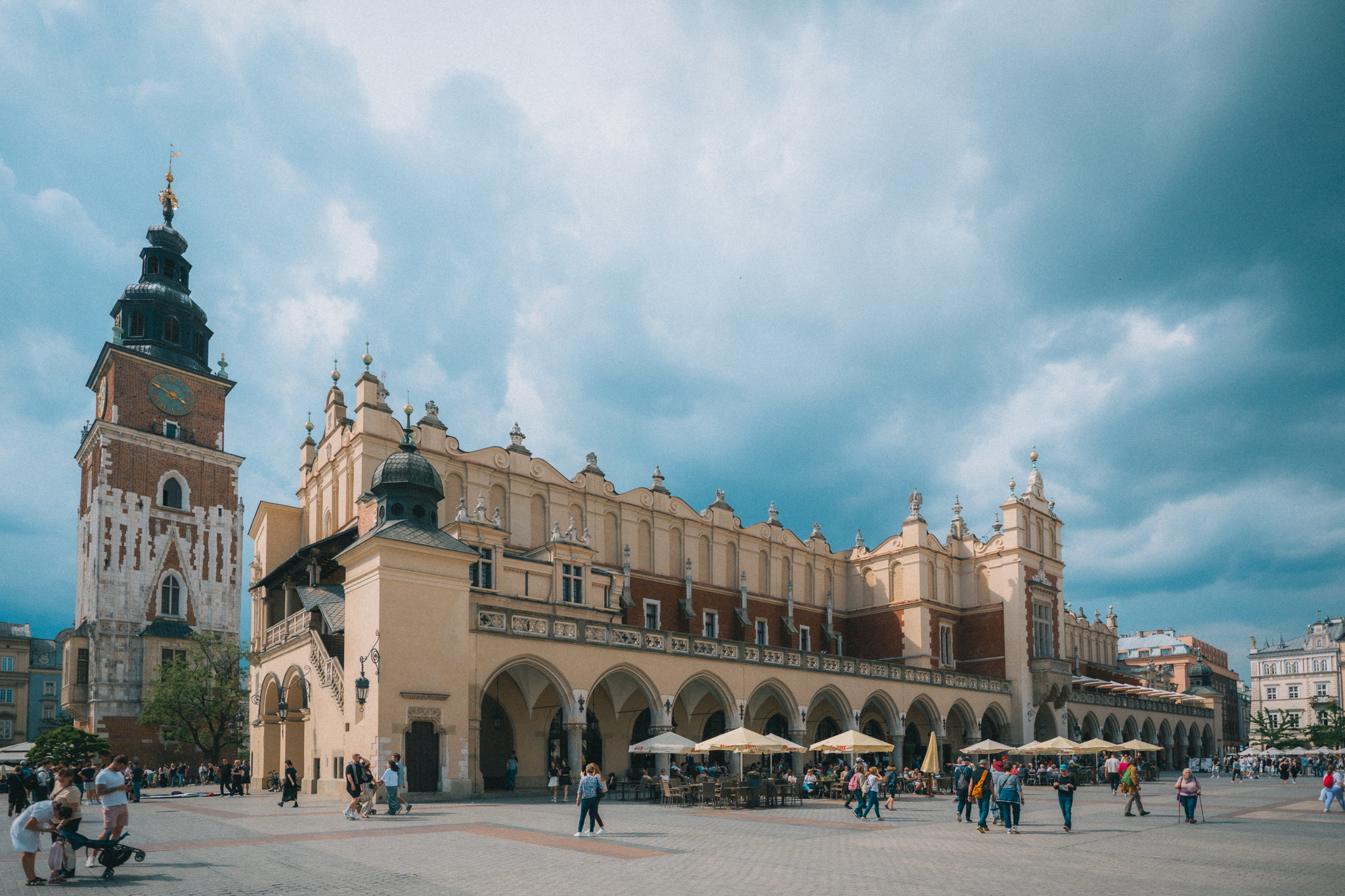 Kraków Main Square (Rynek Główny in Polish). A wide open city square with pedestrians, umbrellas and chairs set up for outdoor dining, and several large historic buildings, one a tall tower and another multi-story longer building that runs the length of the square and houses a market.