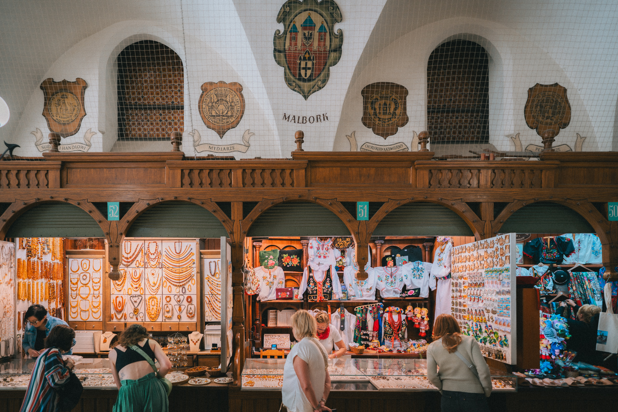 Inside the Cloth Hall in Kraków. Three stalls selling various wares are pictured, from elaborate jewelry to clothing and cloth. The stalls are framed in deep brown wood on the sides and above, with crests lining the white walls above them.