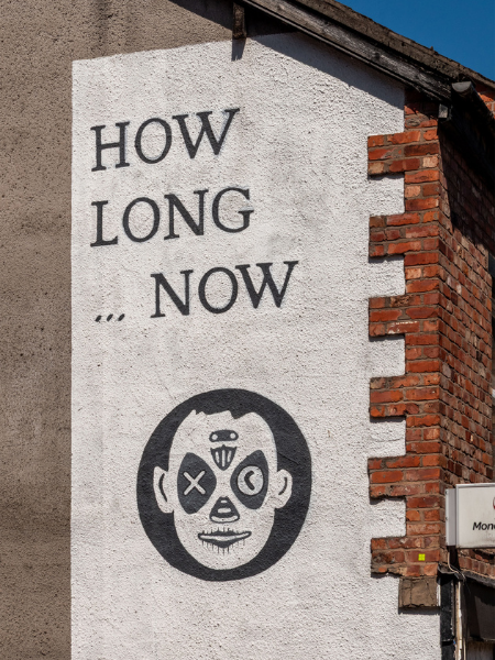 White painted wall in Stockport, UK. Black text: "HOW LONG ... NOW" Below text, black circle with white face inside.