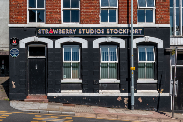 Building facade painted black with all-caps bubble lettering with shading reading "STRAWBERRY STUDIOS STOCKPORT." The "A" in "STRAWBERRY" is replaced by a red strawberry.