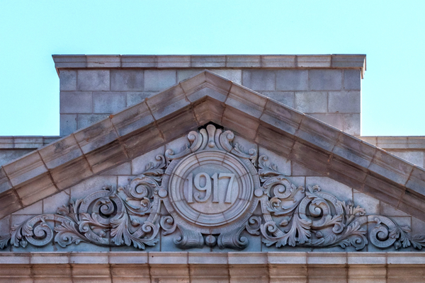 A stone facade with sloping roof and carved triangular panel (pediment) with the year 1917 in a circle surrounded by curved ornamental curls.