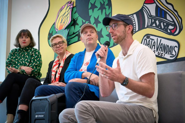 Bill Connolly speaks into a microphone, sitting on stage at the right, with three others on stage to the left.