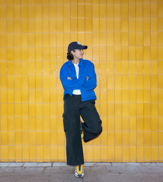 Magdalena Arasanz portrait in a black baseball cap, white shirt, bright blue jacket, loose black pants, and yellow sneakers.