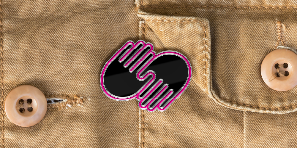 Allkin, symbol of two hands meeting, mocked up as a pin on a khaki button up shirt. Pin: black with bright pink outline.