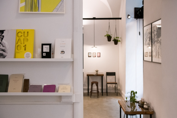Inside the Pressato café. Books on white rail shelves on a white wall. A wooden chair and stool sit around a small wooden table. Photo by Nello Russo.
