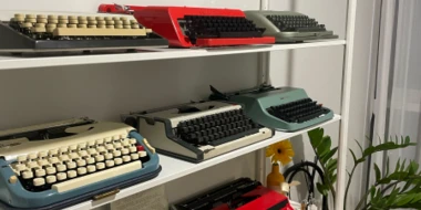 Typewriters on three shelves in a variety of colors (red, teal, grey with white or black keys).