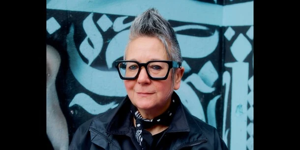 Spike Spondike poses for a headshot in front of a sky blue and black wall covered in designs reminiscent of brush stroke calligraphy. 