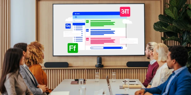 People sit at a meeting room table watching a presentation on a larger screen. The screen contains layered boxes with glyphs and lines suggestive of additional text in a variety of colors (blue, green, red).