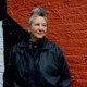 Spike Spondike portrait, wearing a black jacket over a black button-down with a black bandanna tied around her neck, in front of a rust-red wall. Spike Spondike portrait, wearing a black jacket over a black button-down with a black bandanna tied around her neck, in front of a rust-red wall.