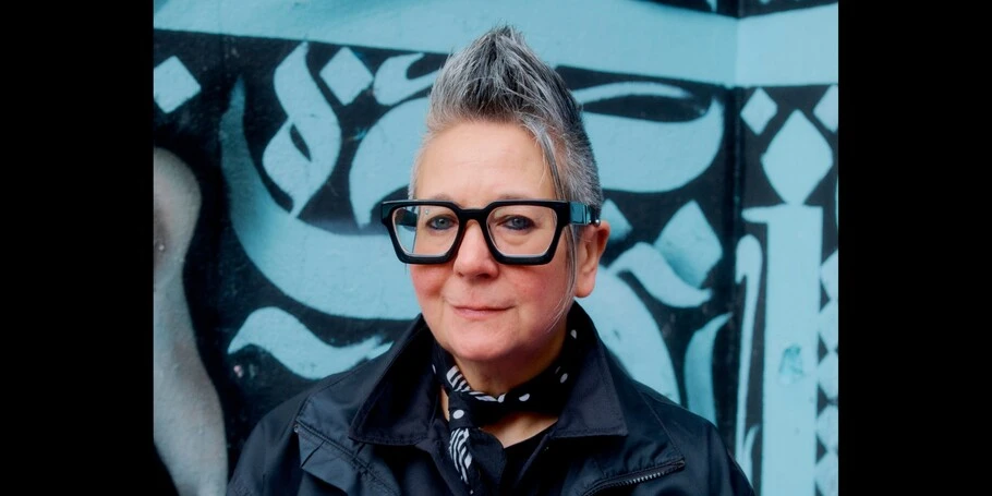 Spike Spondike poses for a headshot in front of a sky blue and black wall covered in designs reminiscent of brush stroke calligraphy. 